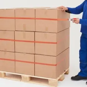 Detalle de la aplicación de cinta adhesiva de fleje con el aplicador manual tesa sobre un palet con cajas de cartón.